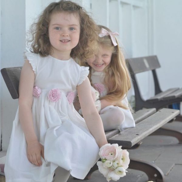 A picture of two flower girls on a bench with white cotton dresses and pink roses - The Little Wedding Company
