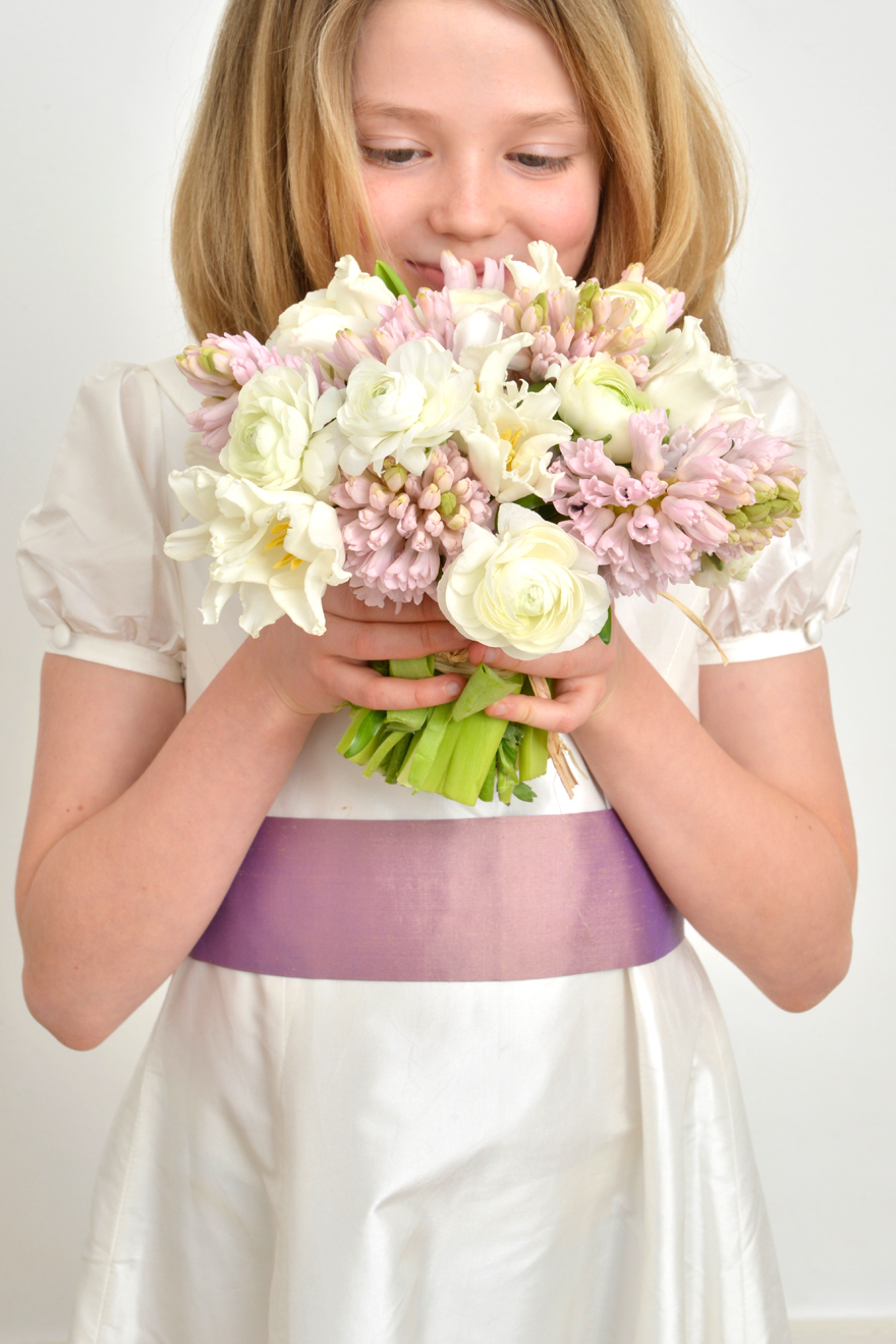 A girl wearing a silk bridesmaid dress with a lilac sash smelling a bridal bouquet - The Little Wedding Company