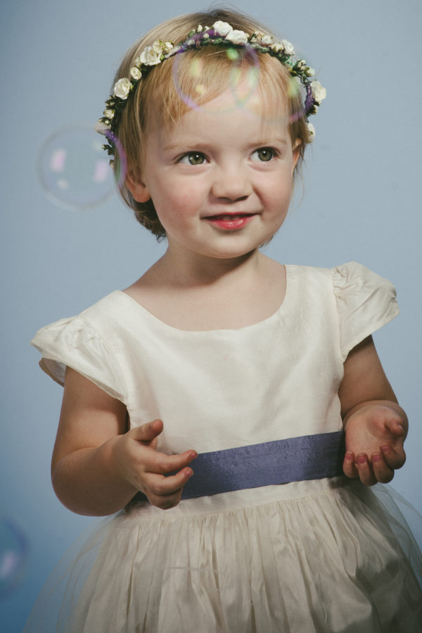 A girl wearing an ivory bridesmaid dress with lavender ribbon and a small floral crown - The Little Wedding Company