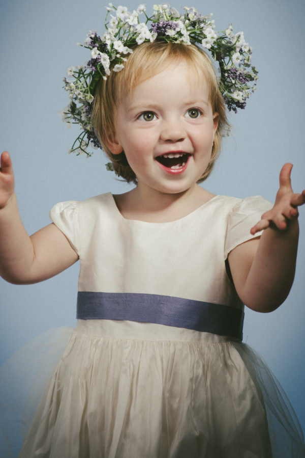 A girl wearing an ivory bridesmaid dress with lavender ribbon and a floral crown - The Little Wedding Company